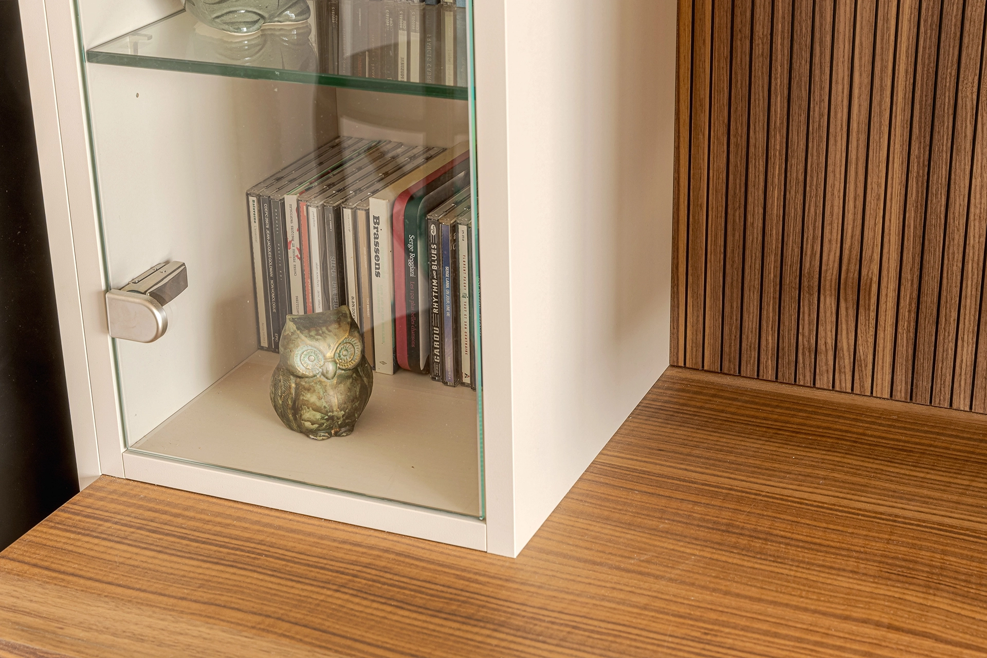 Focus sur les niches vitrées du meuble TV sur mesure, accueillant livres, CD et objets décoratifs, avec assemblage précis entre verre, structure beige et bois à stries verticales. Intérieurs Privés Vannes