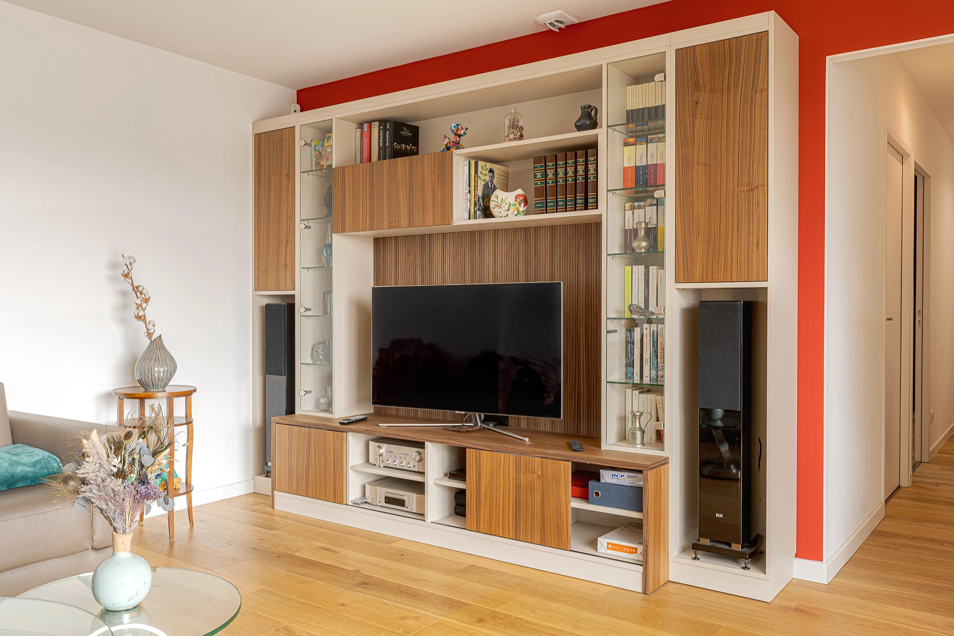 Vue latérale du meuble TV sur mesure occupant tout le mur, combinant façades bois, étagères vitrées, rangement pour enceintes colonnes et panneau central structuré derrière la télévision. Intérieurs Privés Vannes