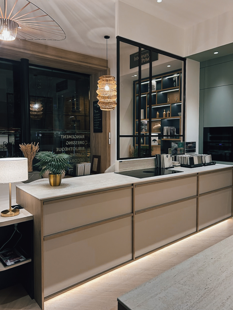 Vue d’ensemble d’une cuisine sur mesure exposée chez Intérieurs Privés Paris 6, associant îlot fonctionnel, rangements bas, verrière atelier et éclairage intégré. Une composition fluide entre cuisine et espace de réception.