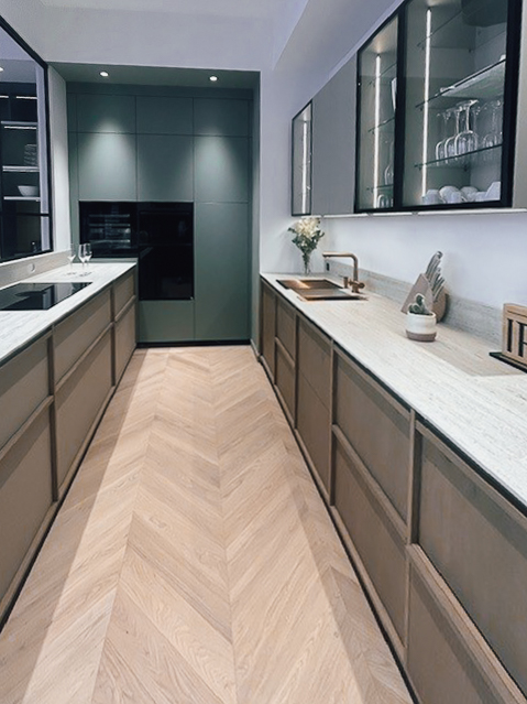 Vue d’ensemble d’une cuisine sur mesure exposée chez Intérieurs Privés Paris 6 : meubles bas et hauts coordonnés, vitrines, crédence minérale et sol en parquet chevron pour une composition sobre et cohérente.