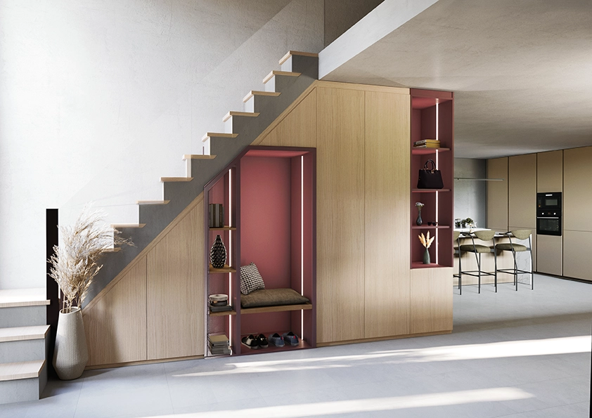 Photo d'un aménagement sous-escalier design et fonctionnel. Il comprend des placards fermés en bois clair, une niche centrale colorée en rose avec un banc confortable et des étagères éclairées, ainsi qu'une bibliothèque latérale. L'ensemble est intégré sous un escalier en béton dans un intérieur moderne avec une cuisine en arrière-plan.