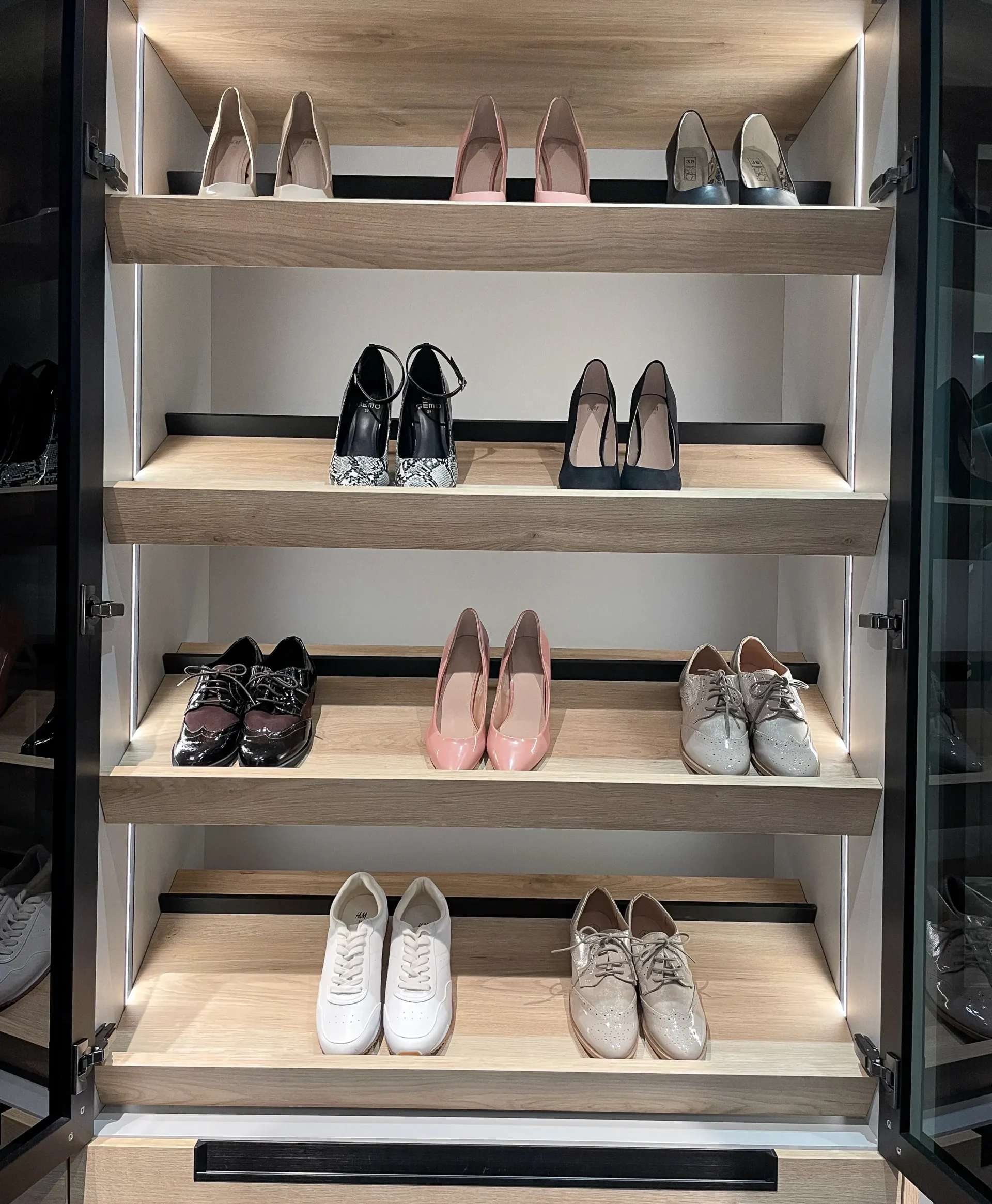 Étagères à chaussures éclairées avec tablettes en bois clair, portes vitrées noires et rangement dédié dans un dressing sur-mesure.