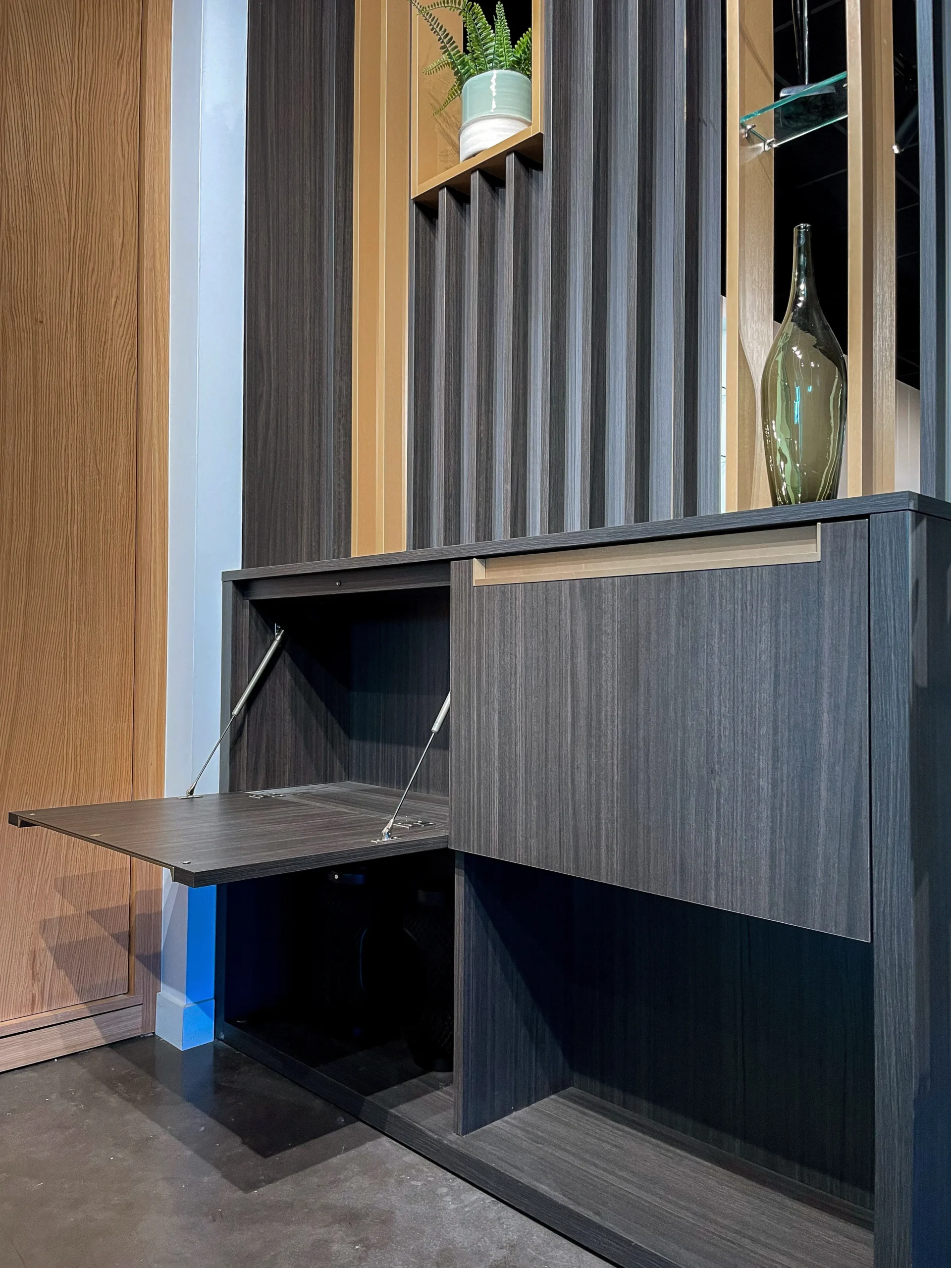 Meuble séparateur sur-mesure avec claustra vertical, niches décoratives, abattant ouvert et finitions bois foncé dans un showroom d’aménagement intérieur.
