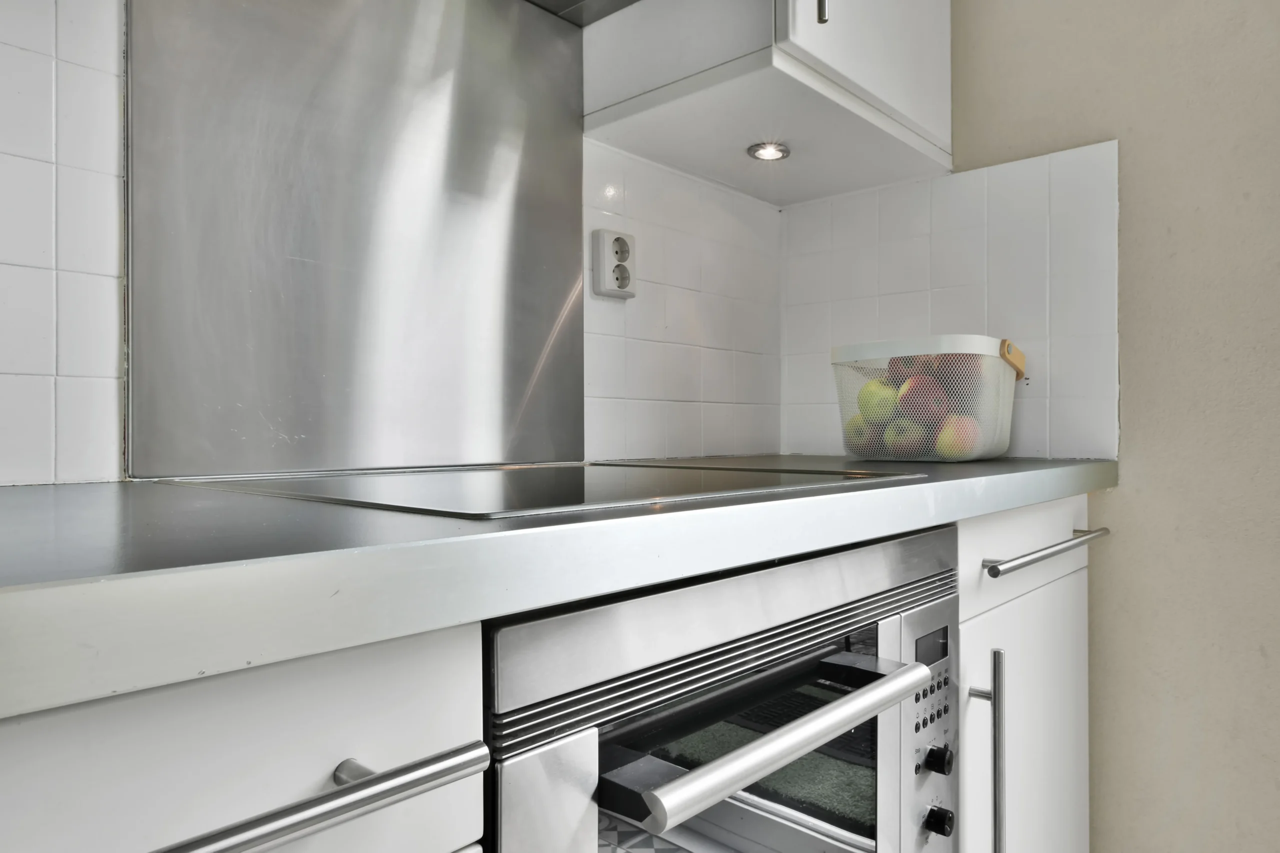 Petite cuisine blanche équipée d’une hotte inox, d’une plaque de cuisson intégrée et d’un four encastré sous le plan de travail.