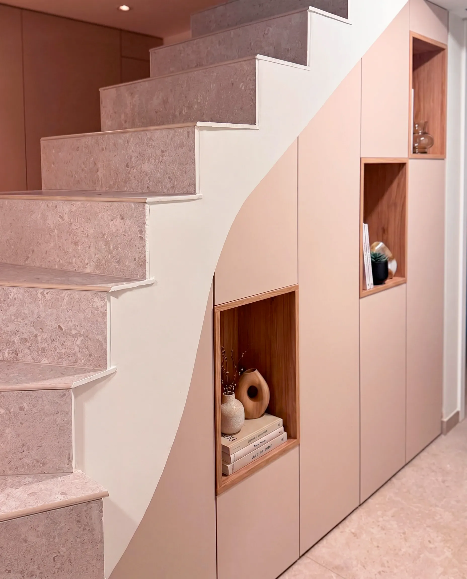 Rangement sous escalier sur-mesure rose poudré avec portes fermées, niches décoratives en bois et optimisation de l’espace sous les marches.