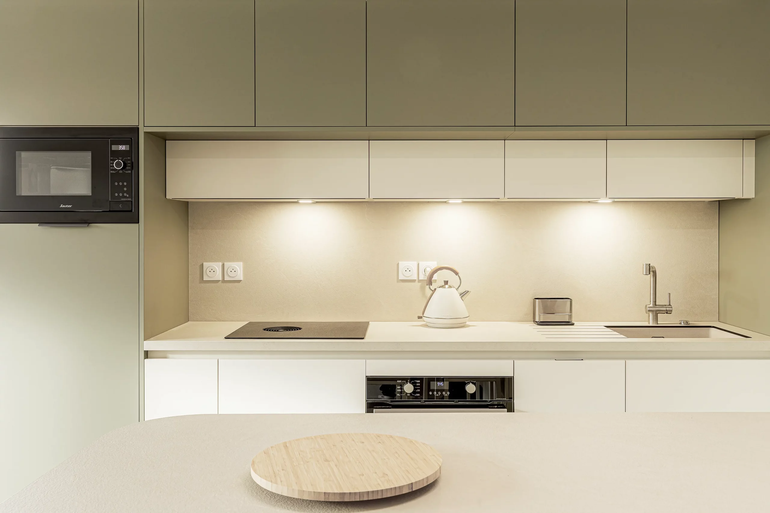 Cuisine linéaire sur mesure avec rangements toute hauteur, façades vert sauge et blanches, plan de travail clair et éclairage intégré