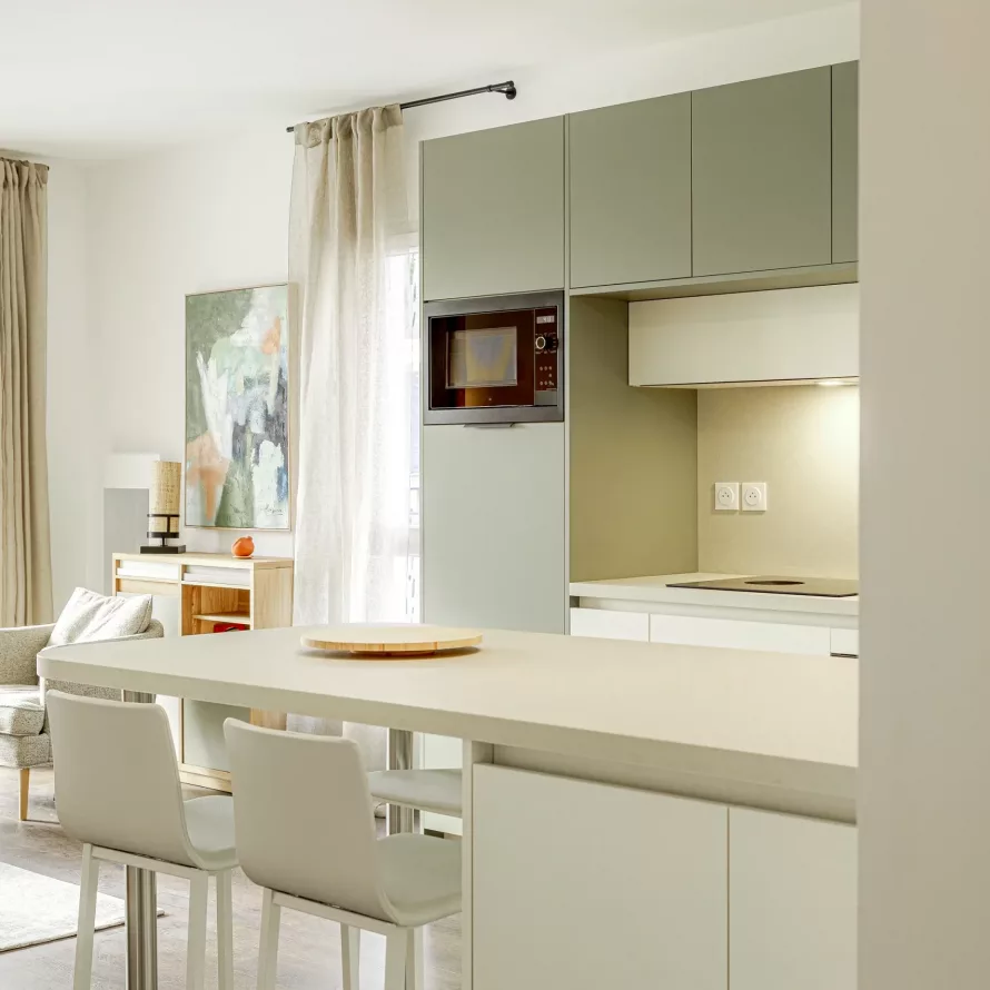 Vue sur une cuisine sur mesure ouverte avec table snack, assises blanches et façades vert sauge