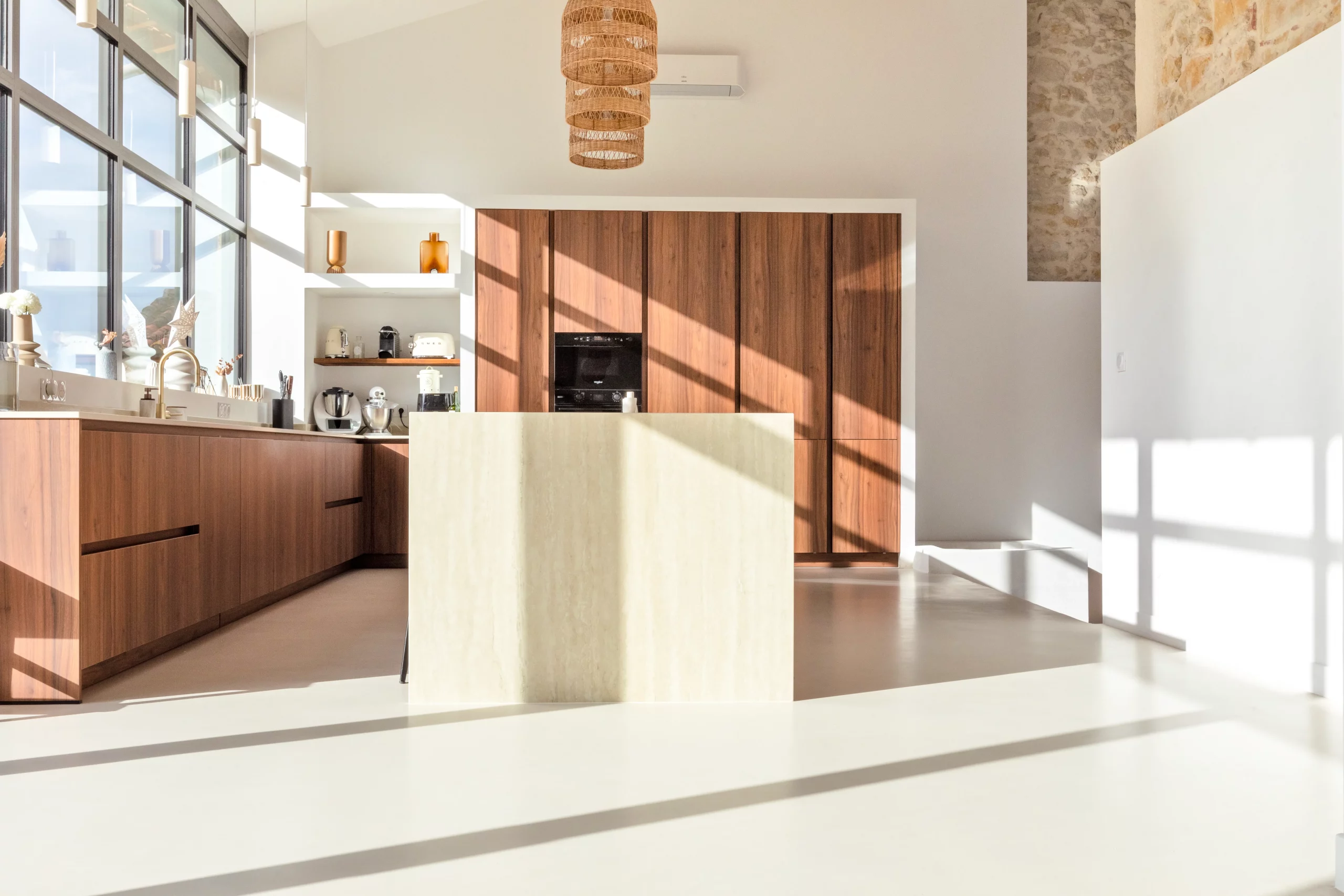 Cuisine avec îlot central clair, rangements aux teintes bois et grandes ouvertures sur l’extérieur