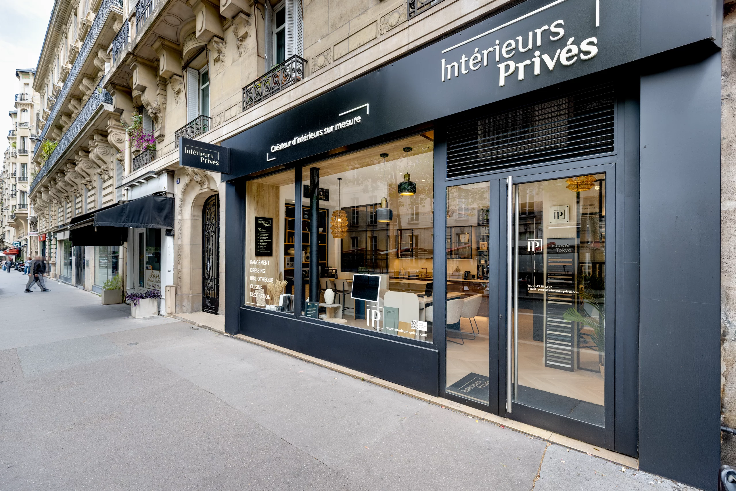 Devanture moderne d'un magasin Intérieurs Privés, créateur d'intérieurs sur mesure, installée au rez-de-chaussée d'un immeuble de style haussmannien. La façade noire élégante dévoile un showroom lumineux à travers de grandes vitrines, mettant en scène du mobilier design.