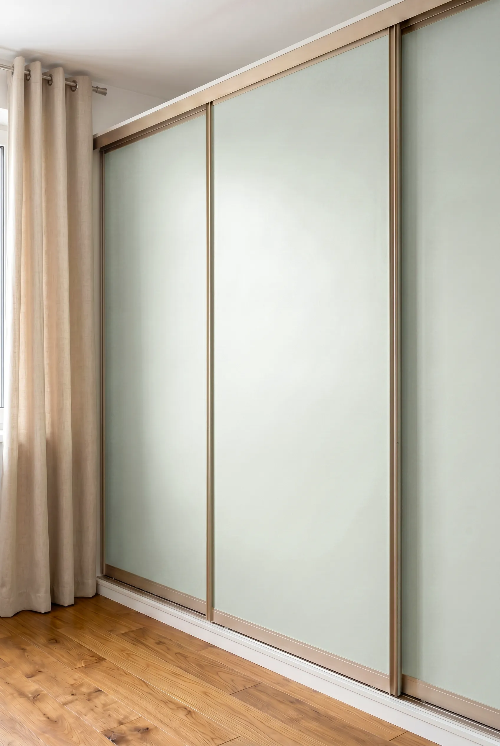 Portes de placard coulissantes sur-mesure vert sauge, avec profils métalliques champagne, intégrées dans une chambre lumineuse avec rideaux beiges et parquet bois.
