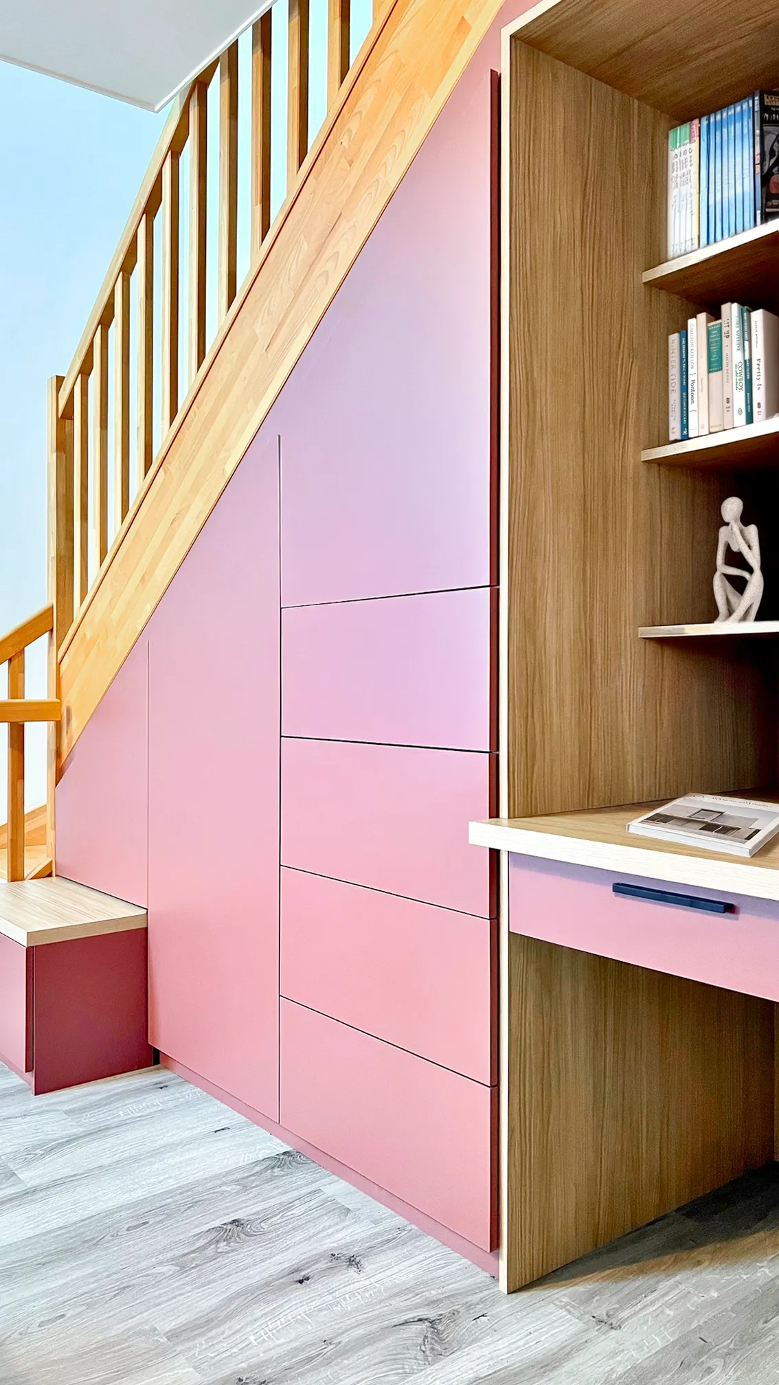 Aménagement sous escalier rose et bois avec placards, tiroirs, niches ouvertes et bureau intégré.
