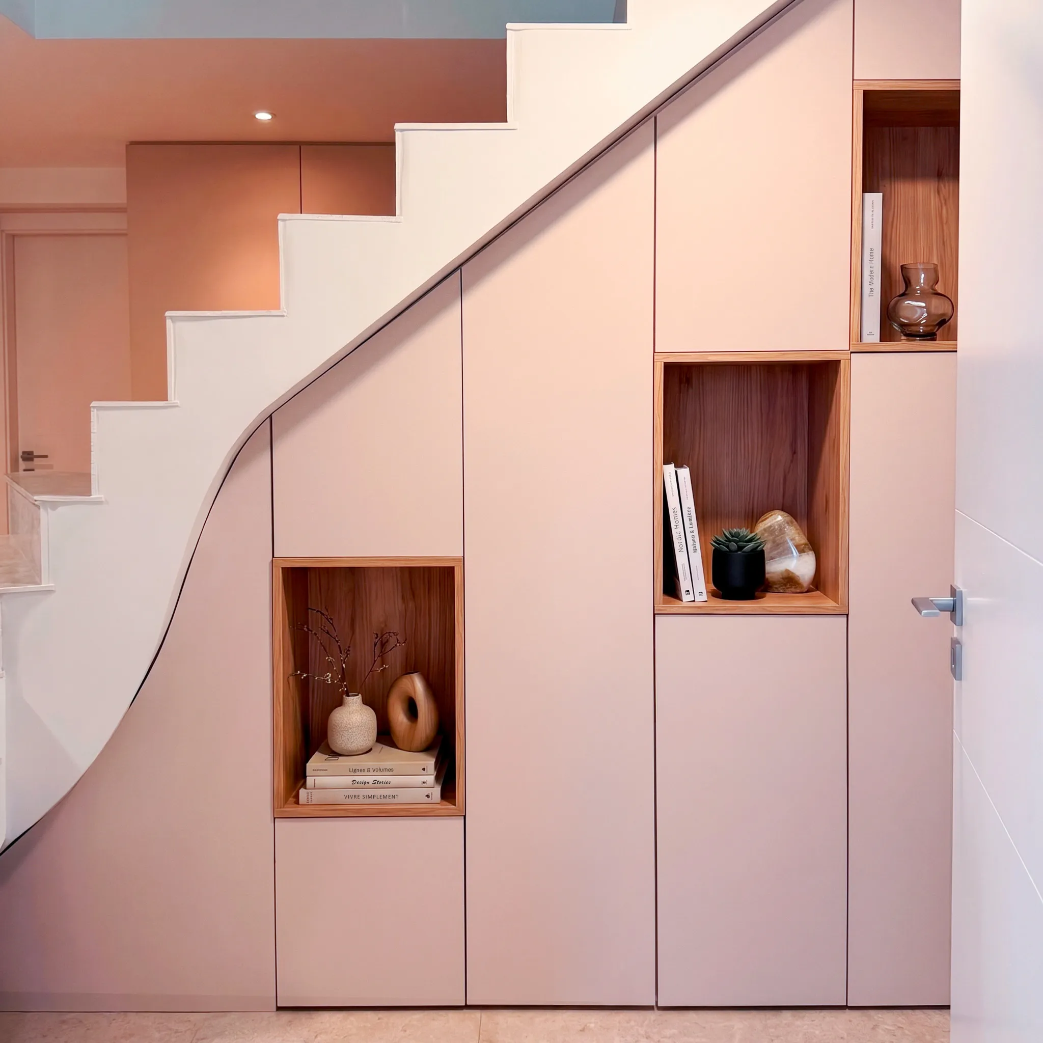 Aménagement sous escalier sur-mesure rose poudré avec façades fermées, niches décoratives en bois et intégration discrète dans l’entrée.