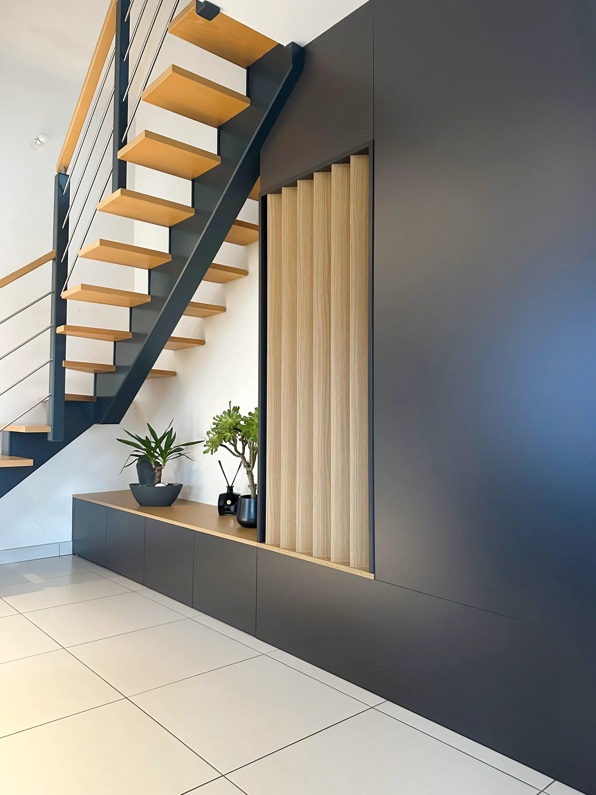 Aménagement sur-mesure installé sous l’escalier avec rangements bas, claustra en bois et façades foncées.