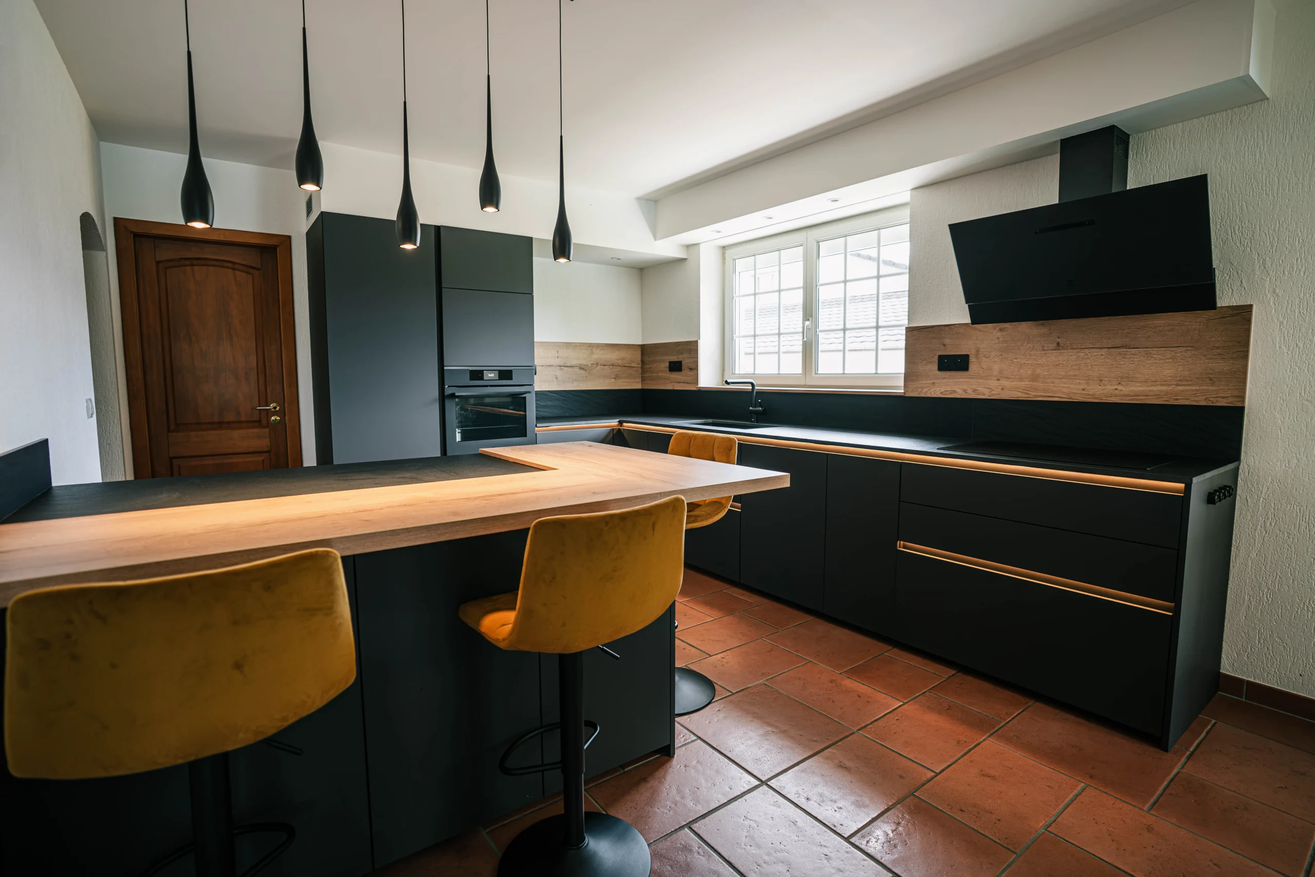 Cuisine sur-mesure à Kembs associant façades noires, finitions bois, plan de travail sombre et espace repas intégré.
