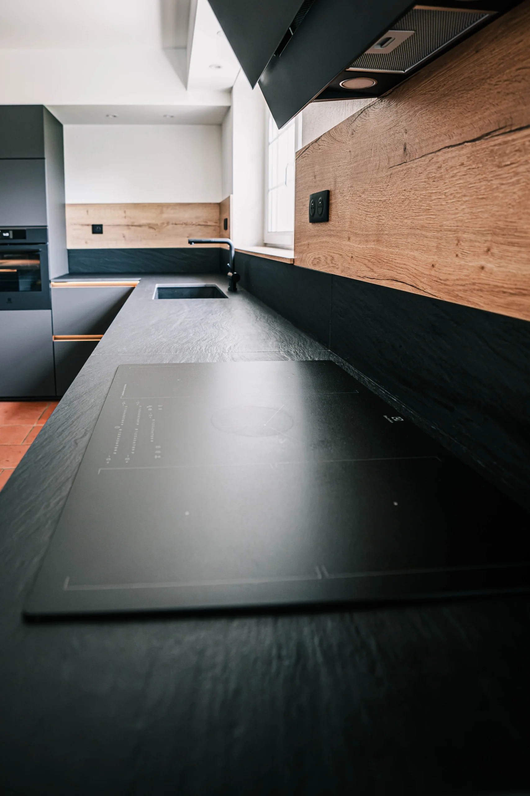 Détail du plan de travail sombre d’une cuisine sur-mesure, associé à une crédence noire et à un habillage décor bois.