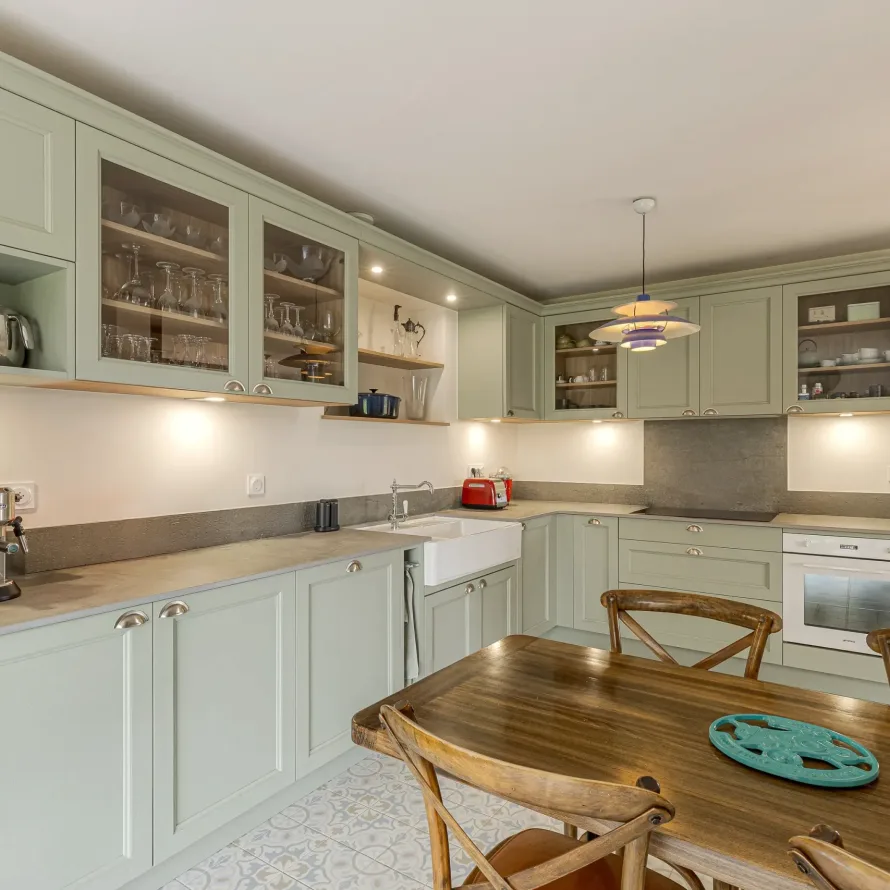 Cuisine sur-mesure en U de style cottage avec façades vert sauge, vitrines, niches ouvertes, plan de travail minéral et coin repas en bois à Vannes.
